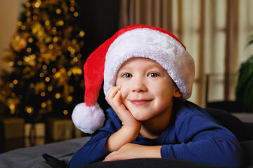 A large portrait of a beautiful boy in a Santa hat on a bed with a gift on the background of a Christmas tree with bokeh lights. The child smiles and looks at the camera.