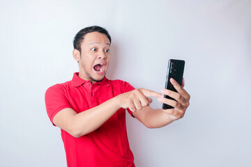 Surprised Asian man wearing red t-shirt pointing at his smartphone, isolated by white background