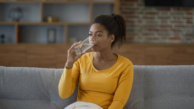 Healthy habits. Positive african american woman drinking water from glass, sitting on sofa at home, free space