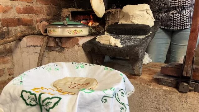 Mujer mexicana torteando maza de maíz en un metate y una estufa de leña para hacer tortillas