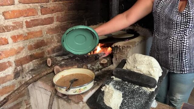 Mujer mexicana torteando maza de maíz en un metate y una estufa de leña para hacer tortillas