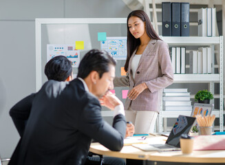 Millennial Asian professional successful female businesswoman presenter lecturer speaker in formal suit standing using pencil pointing graph chart document presentation on glass board in meeting room.