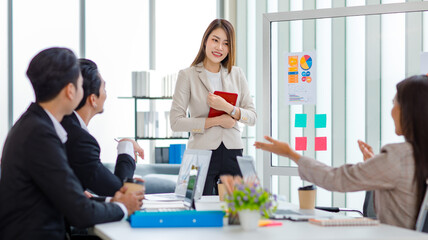 Fototapeta premium Millennial Asian professional successful young cheerful female businesswoman speaker lecturer in formal suit standing smiling pointing graph chart information document on glass board in meeting room
