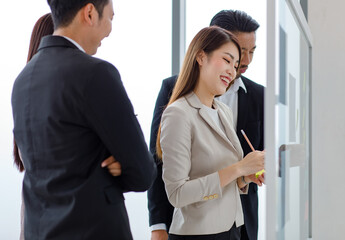 Millennial Asian professional successful female businesswoman presenter lecturer trainer in suit standing pointing paperwork document on glass board presenting to colleagues in meeting room