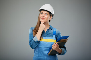 Thinking woman engineer wearing white helmet and overall holding books and clipboard, looking up....