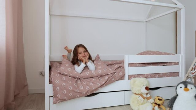 Portrait of a beautiful caucasian little girl smiling while relaxing in her bedroom during a leisure day. Cheerful female kid lying on scandinavian wooden house frame bed.