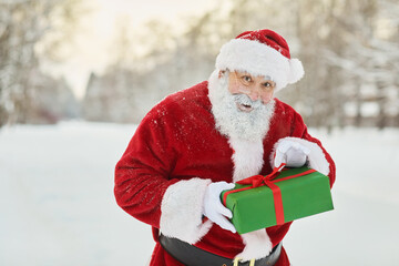 Waist up portrait of traditional Santa Claus holding gift box and looking at camera mischievously in winter forest, copy space