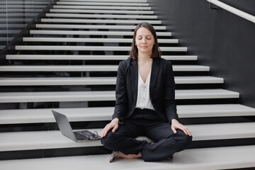 business woman in a formal suit with a laptop in the office of the business center sits on the steps in the lotus position and meditates, relax. Work in a corporation as a marketing financier or