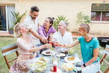 Group of happy middle-aged men and women toasting wine glasses at dinner event at house backyard....