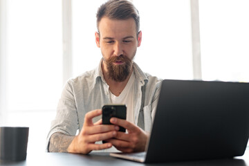 Handsome young bearded man spending time at home with phone in hands.