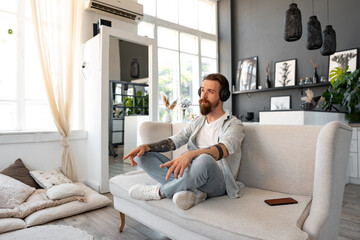 Positive bearded man listening music with headphones while sitting on couch at home