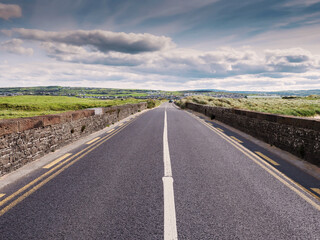 Fototapeta na wymiar Small narrow high quality asphalt road with stone fences on each side. Blue cloudy sky and green fields. Warm sunny day. West of Ireland. Travel concept.