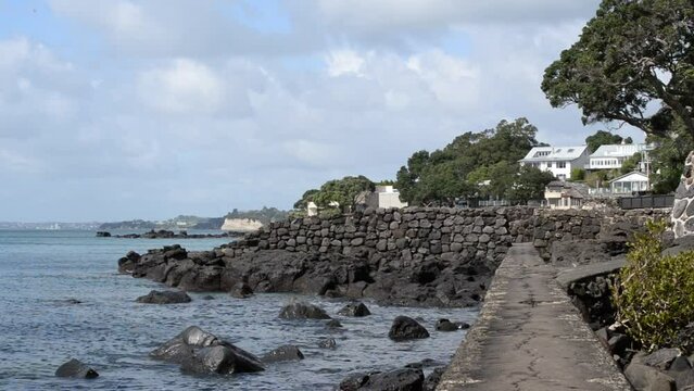 Walking path along the old rocky seawall of a north Auckland beach on a sunny spring day with few cumulus clouds