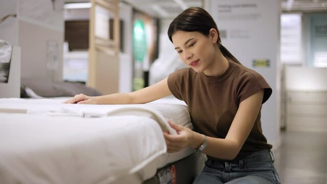 Young beautiful woman choosing mattress at retail furniture store new home. Housewife touching softness test buying bedding for family. Female checking quality shopping bed in mattress store hall.