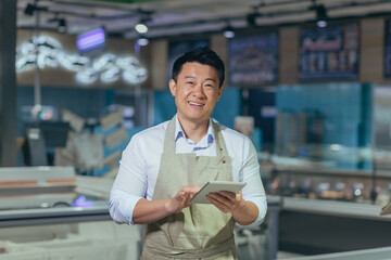 Portrait of a handsome young Asian man, an employee of a store, supermarket. Standing in an apron,...