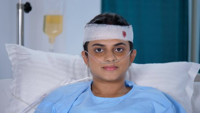 A young Indian woman with a bandage on her head is looking at the camera -accident injury  physical injury  head injury  head wound. A female patient with an oxygen nasal tube resting in the hospit...