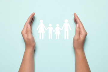 Woman holding hands around paper silhouette of family on light blue background, top view. Insurance...