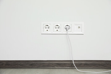 Power sockets with inserted plug on white wall indoors. Electrical supply