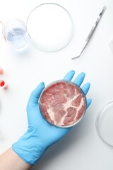 Fototapeta na wymiar Scientist holding Petri dish with cultured meat above white lab table, top view