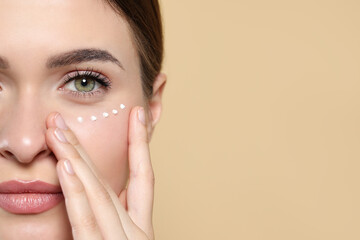 Young woman with cream around eye on beige background, closeup. Space for text