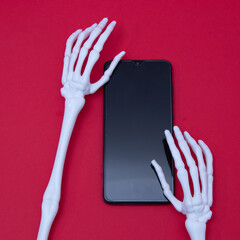 Two skeleton hands holding a mobile phone on a red background. Minimal flat lay Halloween scene.