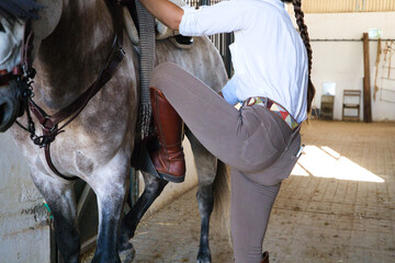 Young and beautiful woman gets on a Thoroughbred horse for the competition. The woman is in the stable where she prepares the Spanish thoroughbred horse. Detail of the foot in the stirrup.