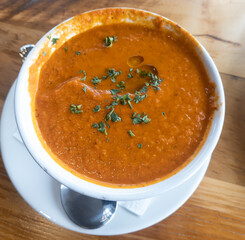 A rich and creamy tomato soup with drops of extra virgin olive oil topped with parsley. The soup is in a white bowl with a spoon on the side on a wooden table. 