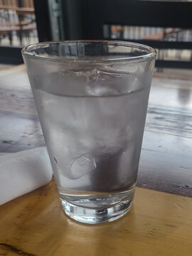 A glass of water served at a resturaunt onto a wooden table, the silverware wrapped in a naplin is to the left side of the glass.