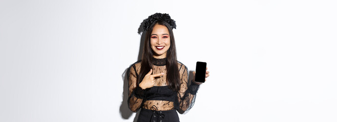 Happy beautiful asian girl in witch costume pointing finger at smartphone screen with pleased...