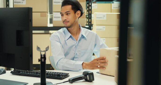 Happy Asian man wear formal uniform shirt sit in front of desk use computer and barcode machine scanner record customer online data in cardboard box delivery at warehouse. Startup business concept.
