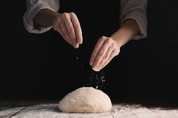 Woman sprinkling flour over dough at wooden table on dark background, closeup