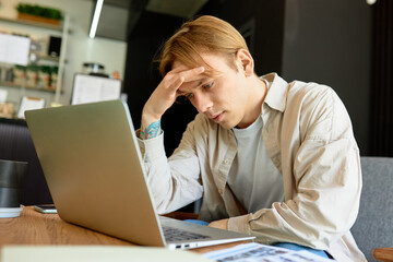 Portrait of tired upset exhausted man sitting in front of laptop with hand over forehead, having headache, trying to concentrate and read information on screen. Freelance and deadline