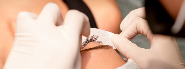 Fototapeta na wymiar The hands of the cosmetologist are gluing white tape under the eye of the young caucasian woman during the eyelash extension procedure, closeup