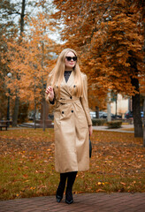  portrait of blonde woman in sunglasses  and in beige coat walking  in autumn park