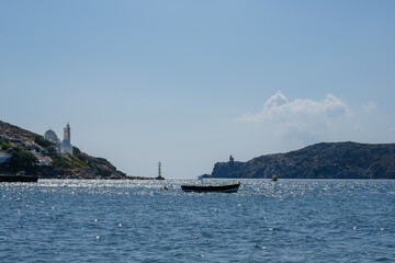 Obraz premium View of a little fishing boat with a Greek flag, a beautiful church and the lighthouse in the background at the port of Mylopotas in Ios Greece