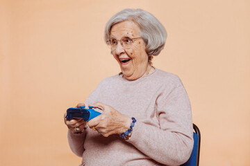 Close up portrait of excited elder grandmother with toothy smile playing video game at studio isolated over beige background. Player, console, gamer concept. © carlesmiro