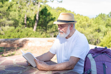 older hiker man sitting at an outdoor table among the trees with reading a book