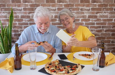 Happy senior couple having fun together with pizza and beer. Elderly woman and man laughing sitting at home table using phone for a selfie