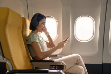 Young woman with mobile phone and headphones listening to music in airplane during flight