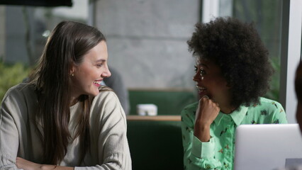 Diverse women at coffee shop in conversation. Three group of girlfriends speaking seated at cafe place with laptop laughing