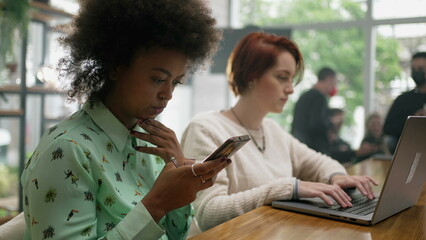 Fototapeta premium People using cellphone and laptop at coffee shop. Two young women staring at screens. Girl looking at computer browsing internet inside cafe restaurant