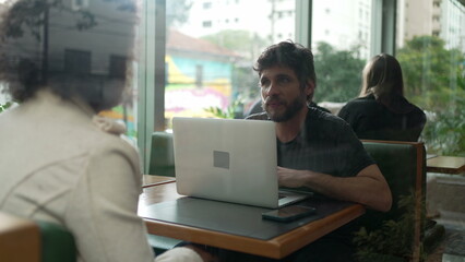 People inside coffee shop through glass window. Men and women customers talking inside cafe restaurant. Person in front of laptop chatting with female friend