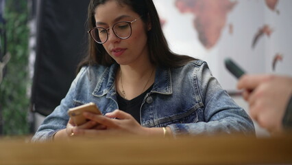 Candid young woman using phone seated at coffee shop. South American Girl in 20s staring at smartphone device