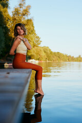 A middle-aged woman is relaxing on the dock and doing yoga breathing exercises. She put her feet in...