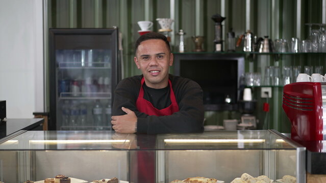 Barista leaning on counter smiling at camera. Portrait of a hispanic employee wearing red apron behind coffee shop counter. Small local business store