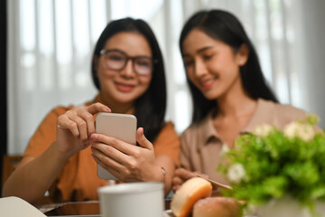 Two happy young woman using smart phone while sitting in modern coffee shop