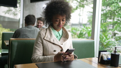 One black woman checking smartphone device sitting at coffee shop table. An African American girl browsing internet on phone