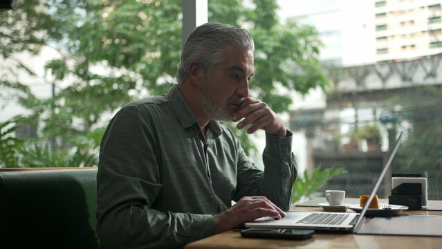 Middle Aged Man Working At Coffee Shop In Front Of Laptop. Older Person Receiving Message Notification On Phone. Executive Person Typing On Computer