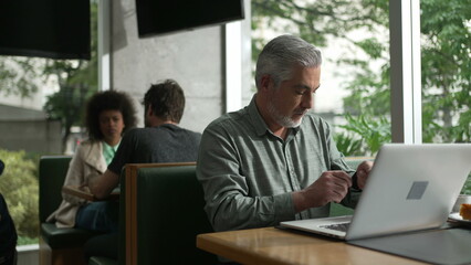 Fototapeta premium Middle aged man preparing to work adjusting shirt and organizing coat and items in front of laptop seated at coffee shop