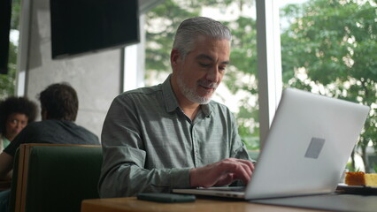 Fototapeta premium Middle aged entrepreneur working at coffee shop in front of laptop. Senior male executive typing on computer keyboard checking phone and using modern technology